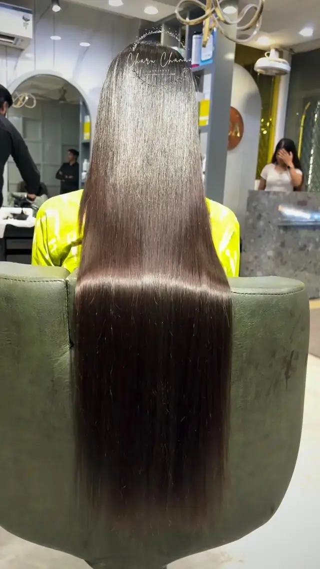 A woman seen from behind with very long, straight, glossy dark hair reaching her waist, seated in a salon chair, a stylist standing beside her.