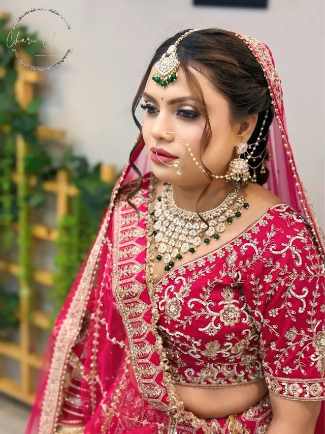A bride in a pink and gold lehenga with a green and gold maang tikka and a nose ring joined by a chain, standing in front of a wall of greenery.