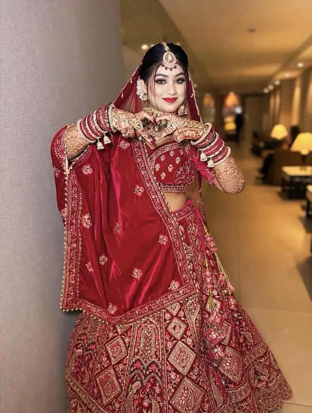 A bride in a red embroidered lehenga with a gold maang tikka and heavy chooda, one hand raised, standing in a lit salon corridor.