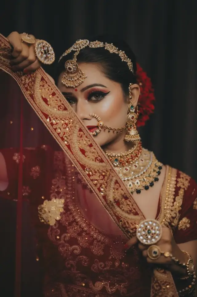 A close-up of a bride in maroon and gold with a heavy layered necklace, a large maang tikka and a nose ring, one bangled arm raised across the frame.