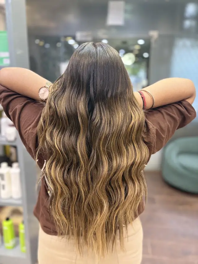 The back of a woman's long wavy hair coloured in caramel balayage, her hands lifted through it, inside the salon.