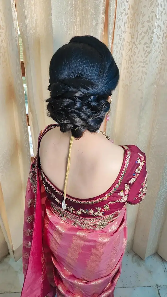 The back of a bride's low twisted bun with a maang tikka chain running across it, over a pink embroidered blouse.
